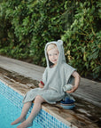 A smiling child in a mushie gray hooded towel plays with Bath Boats by the pool.