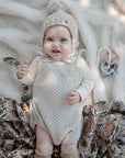 Baby on mushie Organic Cotton Muslin Swaddle Blanket, wearing knit outfit and hat with toys nearby.
