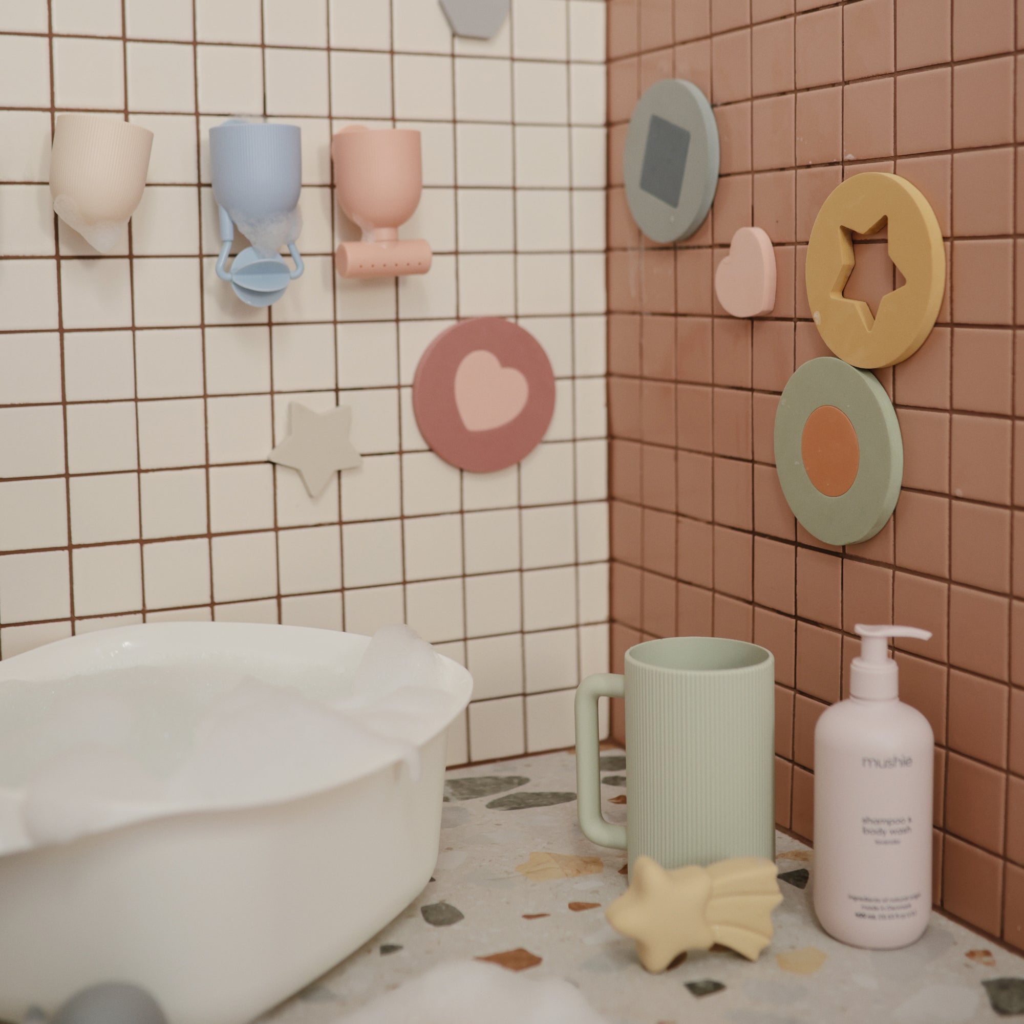 Mushie Baby Shampoo &amp; Body Wash beside children’s bath toys and cups on a tiled ledge.