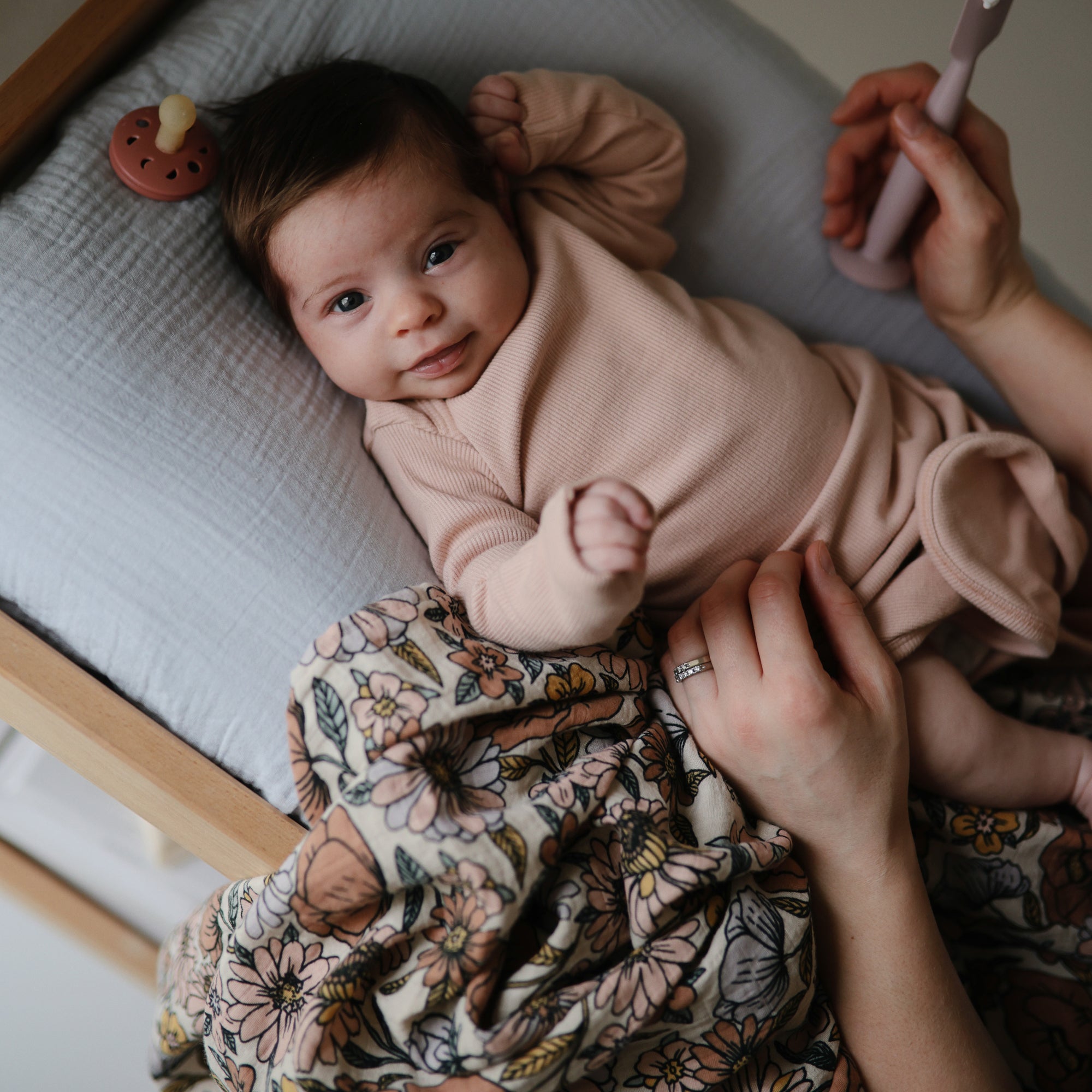 Baby in pink on pillow with mushie Organic Cotton Muslin Swaddle Blanket and floral blanket nearby.