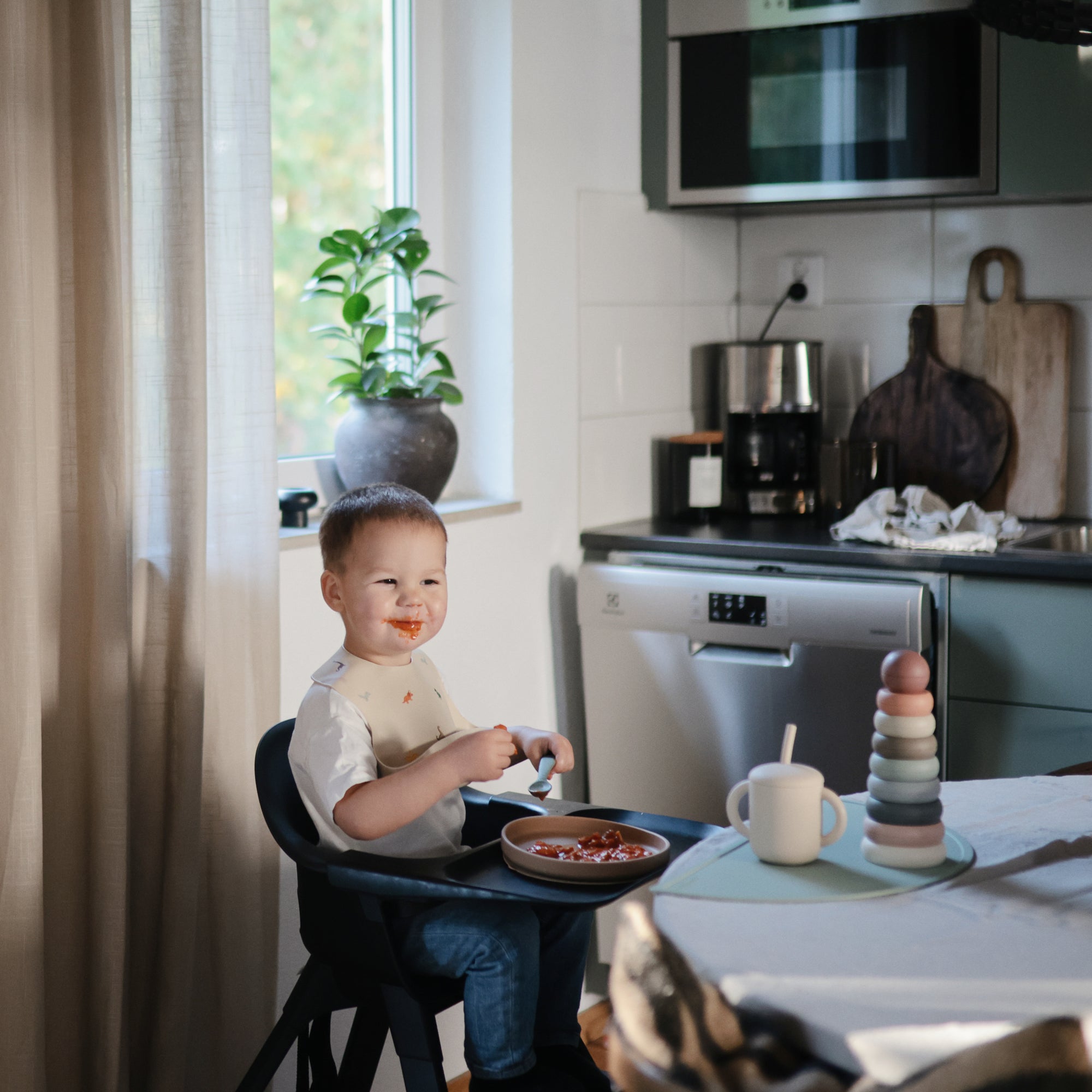 Smiling toddler in a high chair wears a mushie Silicone Baby Bib in a cozy kitchen.