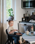 Smiling toddler in a high chair wears a mushie Silicone Baby Bib in a cozy kitchen.