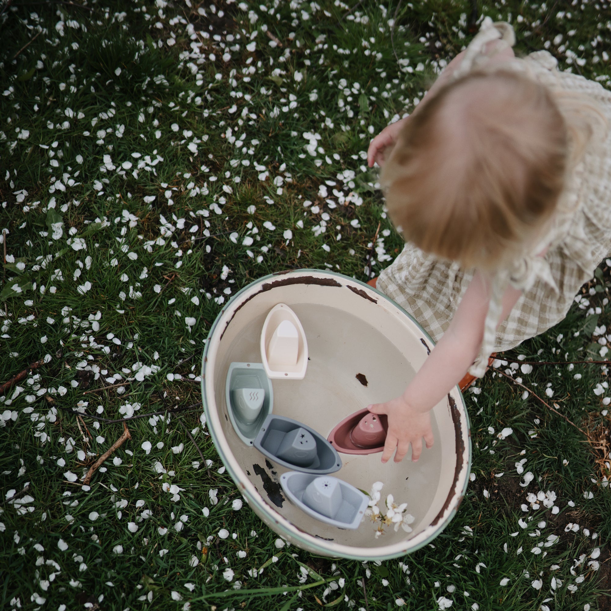 A toddler plays with mushie Bath Boats on grass and flower petals.