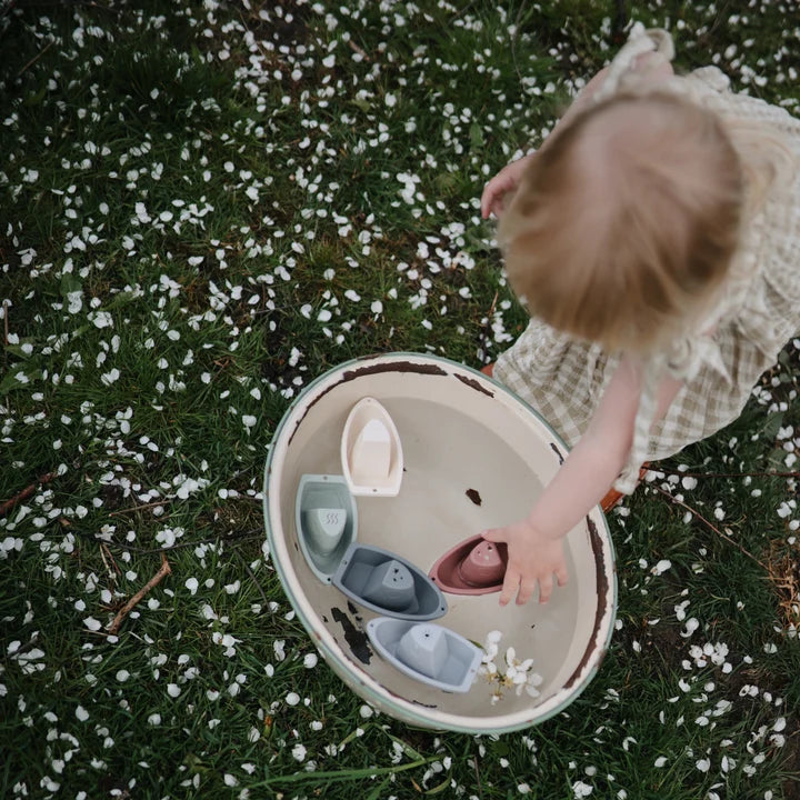 Child playing with toy boats in a bowl outdoors on grass with small white flowers.
