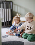 Two young children play with mushie’s Stacking Cups Toy on a rug in a cozy nursery room.