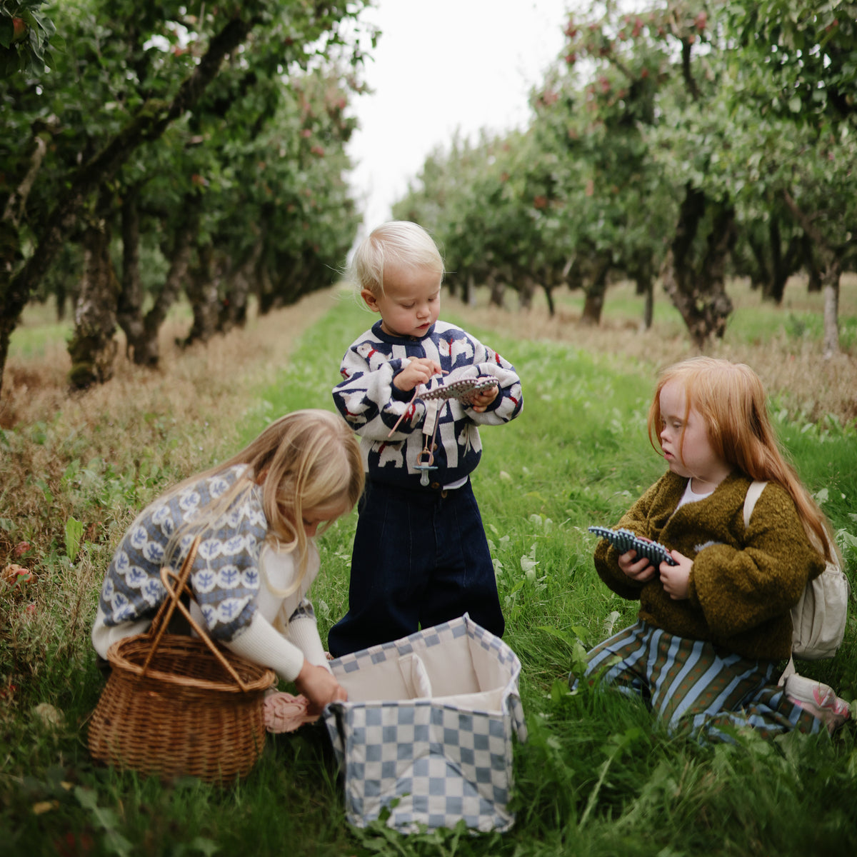 Three kids pick apples in an orchard, filling baskets and a mushie Diaper Caddy.