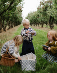 Three kids pick apples in an orchard, filling baskets and a mushie Diaper Caddy.