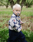 Toddler with FRIGG Daisy Silicone Pacifier holds a notepad in an orchard with fallen apples.