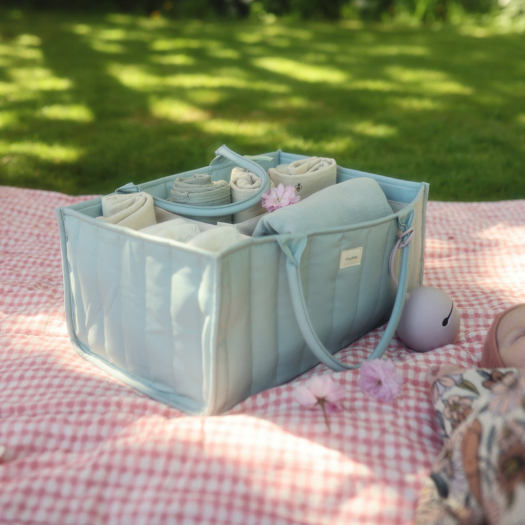 Light blue organizer bag with rolled towels and mushie Silicone Pacifier Case on a picnic blanket.