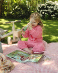 Young girl in pink pajamas plays with mushie Reusable Sticker Set on a picnic blanket.
