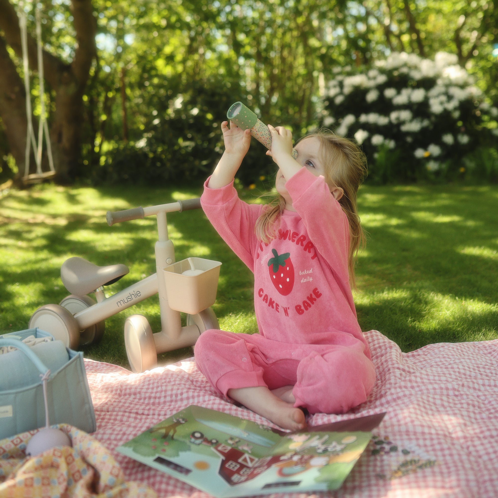 Young child in pink pajamas beside a mushie Mini Toddler Bike looks through a toy tube outdoors.