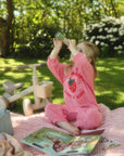 Young child in pink pajamas beside a mushie Mini Toddler Bike looks through a toy tube outdoors.