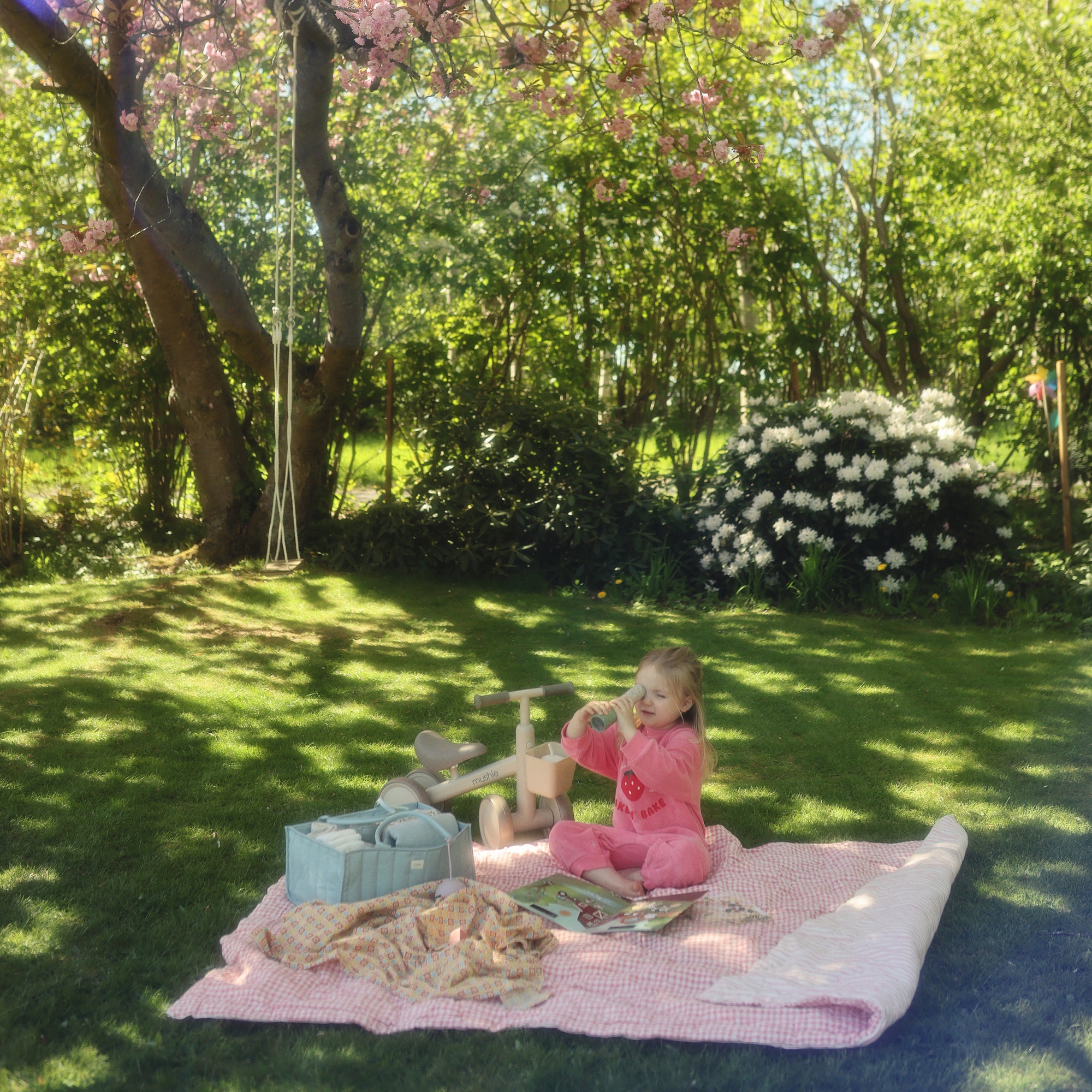 A young girl sits on a picnic blanket beside a mushie Diaper Caddy in a sunny garden.