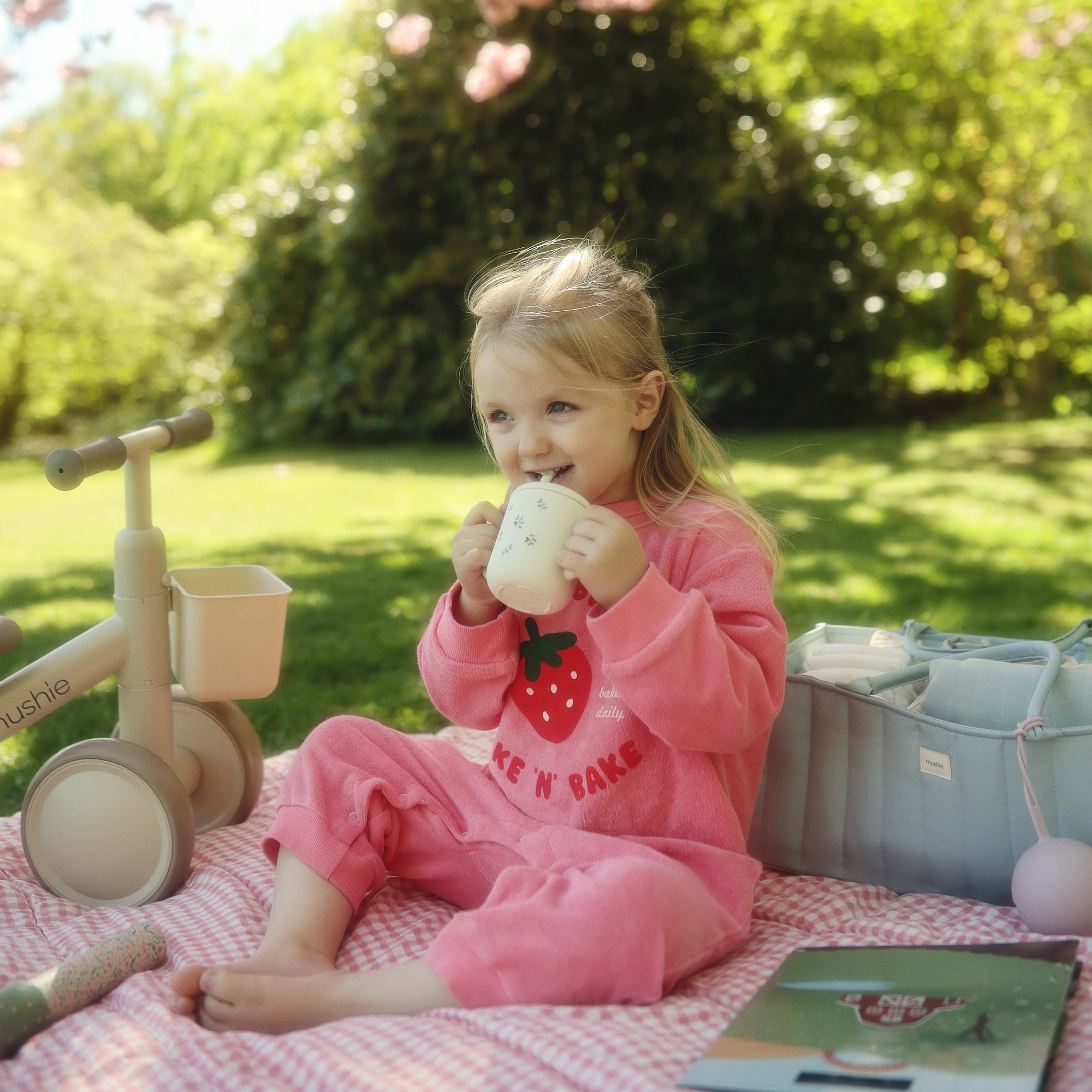 A young child drinks from a cup near a mushie Diaper Caddy outdoors.