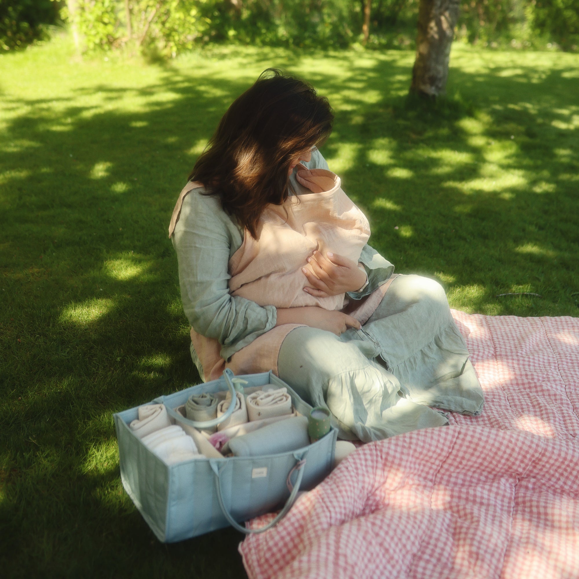 Woman on grass breastfeeding near a picnic blanket and a mushie Diaper Caddy.