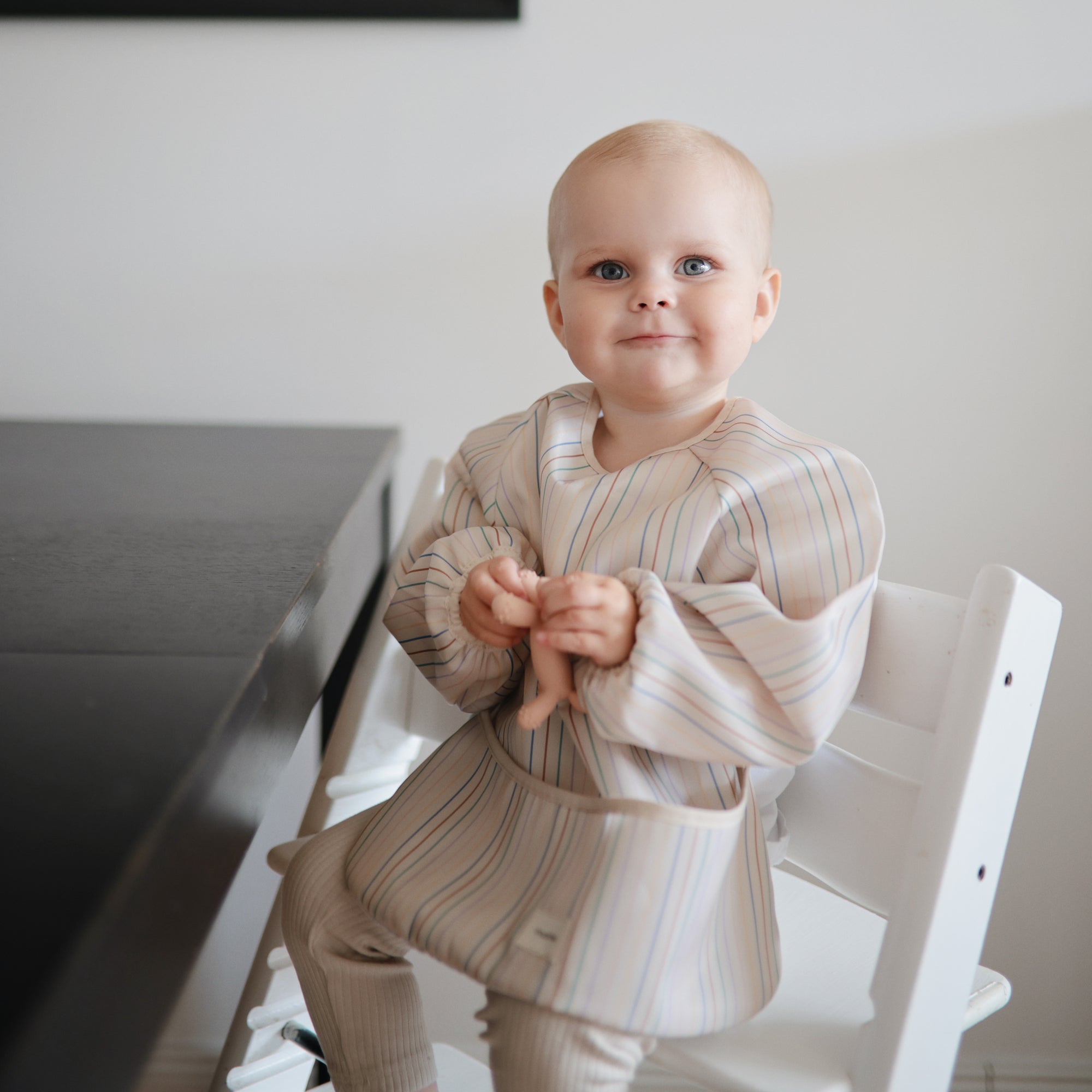 A baby in a striped bib holds a mushie Figurine Teether on a white chair by a dark table.