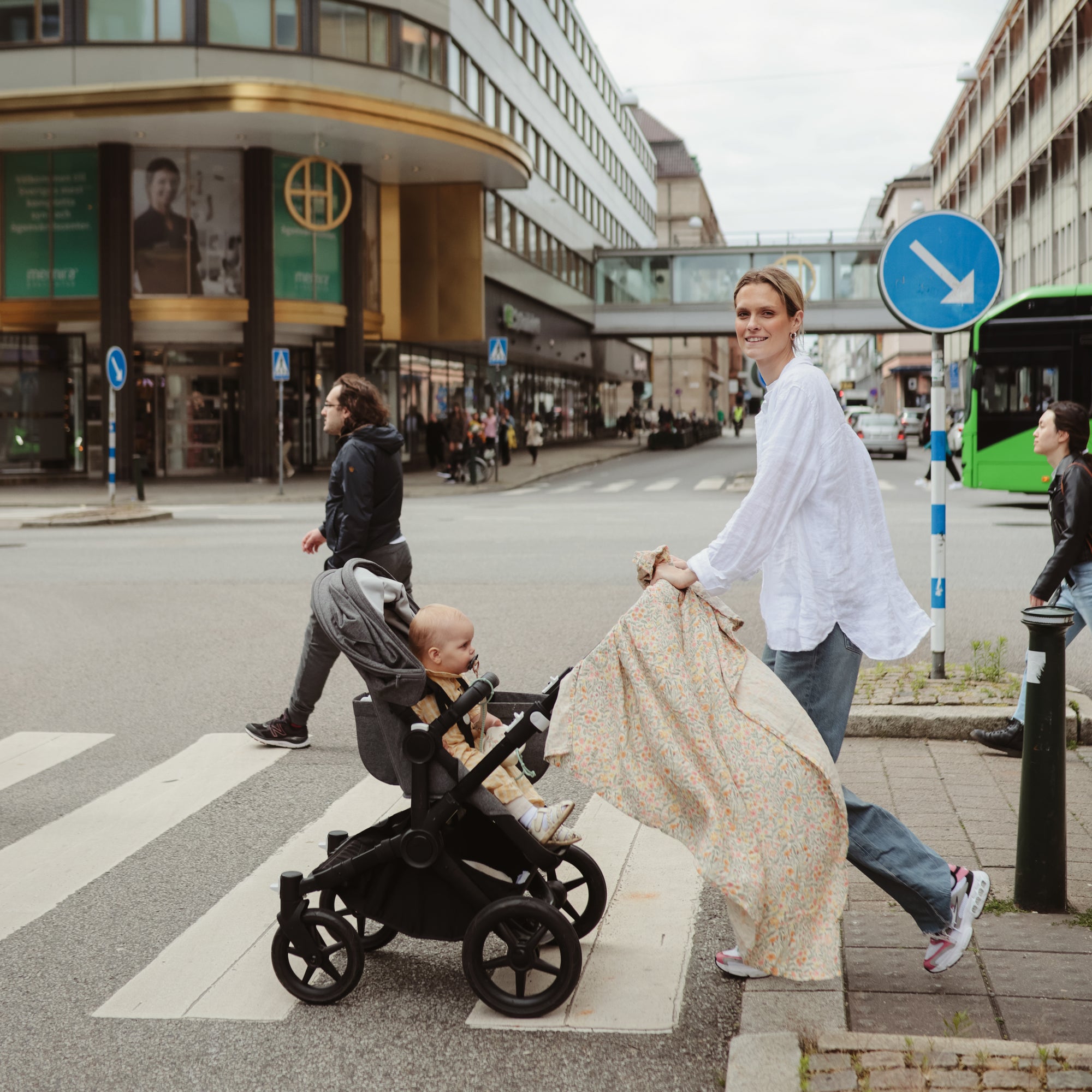 Smiling woman pushes stroller; baby wrapped in mushie Organic Cotton Muslin Swaddle Blanket.
