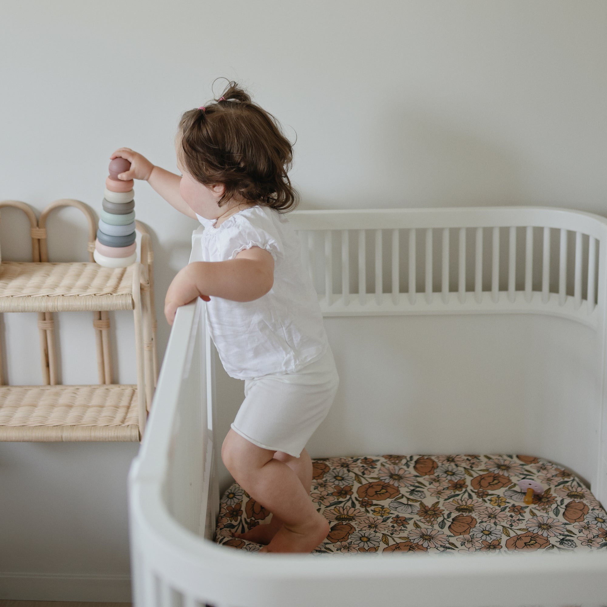 Toddler in crib on mushie Extra Soft Muslin Crib Sheet, holding rings and facing a shelf.