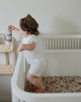 Toddler in crib on mushie Extra Soft Muslin Crib Sheet, holding rings and facing a shelf.