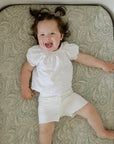Smiling toddler on mushie Extra Soft Muslin Crib Sheet, wearing white clothes.