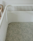 Mushie Extra Soft Muslin Crib Sheet on white crib next to wicker shelf and stacked rings toy.