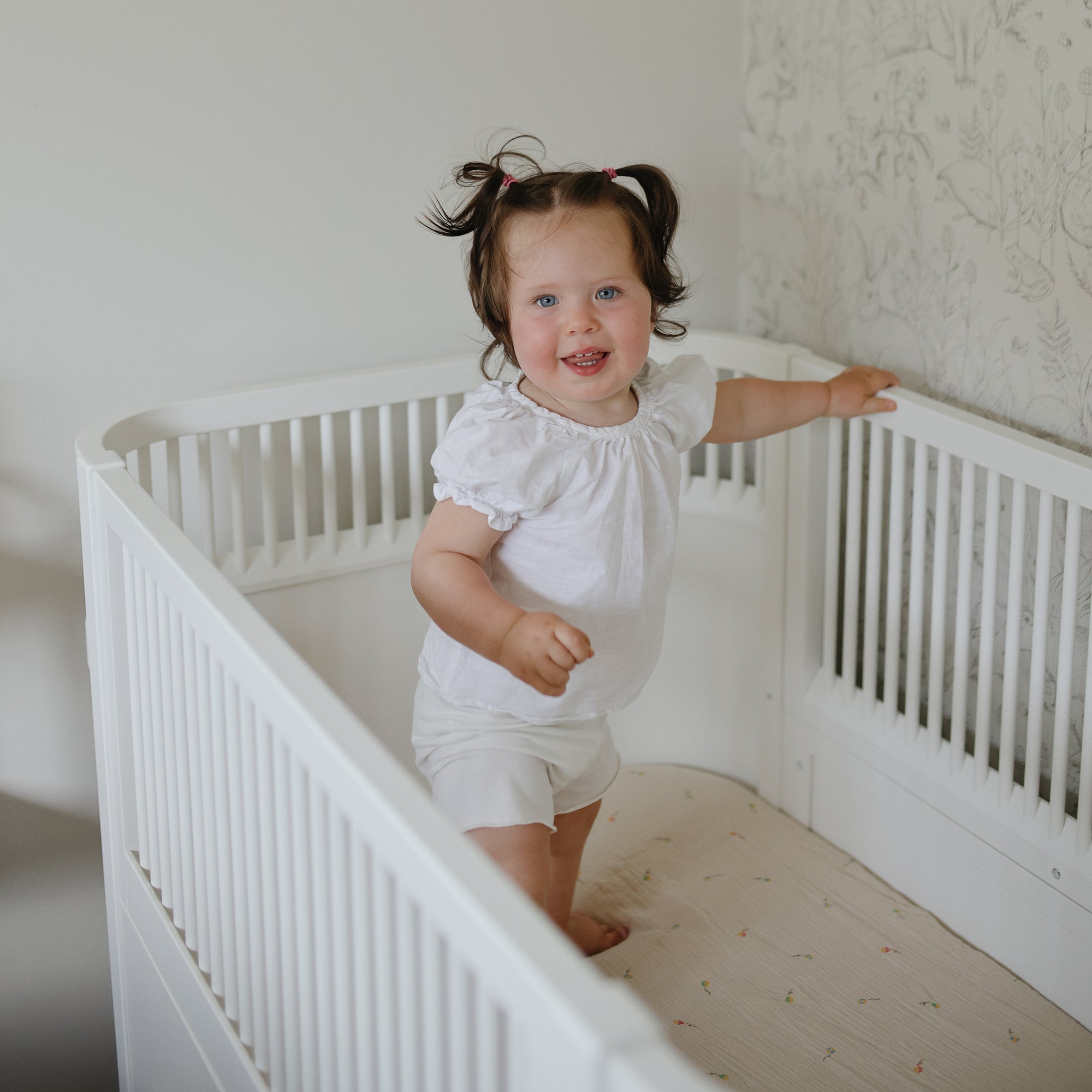 Toddler smiles in white crib on mushie Extra Soft Muslin Crib Sheet, holding the rail.