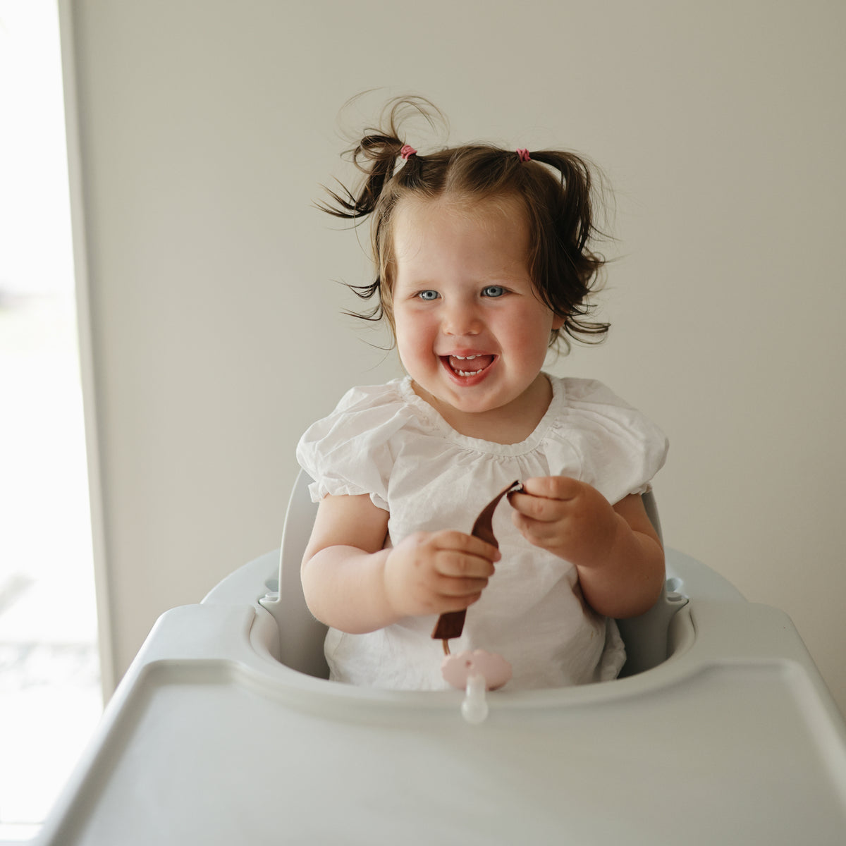 Smiling toddler in high chair holds food and a mushie Linen Pacifier Clip 2-Pack.