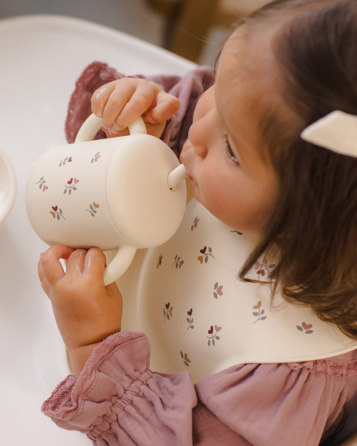 Toddler uses Rylee + Cru Feeding Kit Heart Ditsy bib, sipping from cup in highchair.