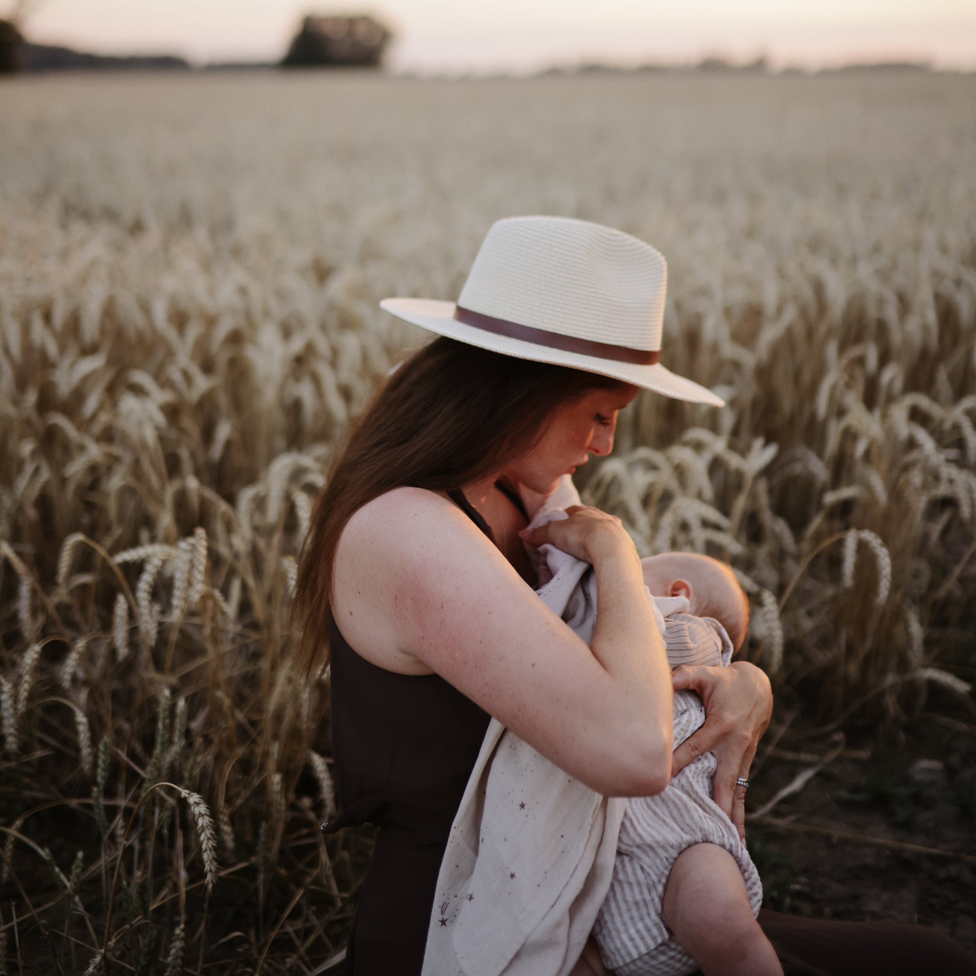Woman in a hat nursing with mushie Organic Cotton Muslin Swaddle Blanket at sunset in wheat field.