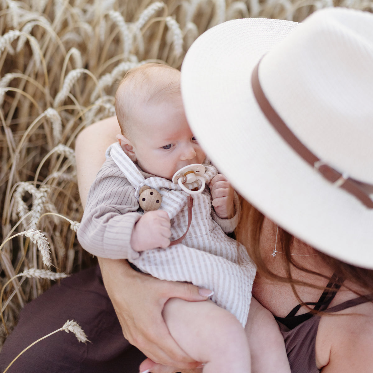 Woman in hat holds baby with mushie Linen Pacifier Clip 2-Pack in field of tall wheat grass.
