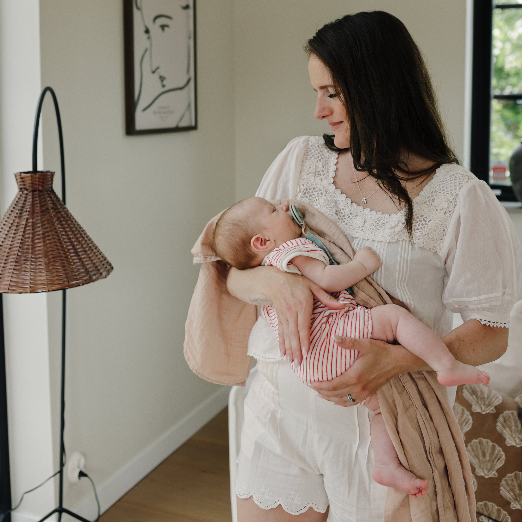 Woman in white holding a baby wrapped in mushie Organic Cotton Muslin Swaddle Blanket, cozy room.