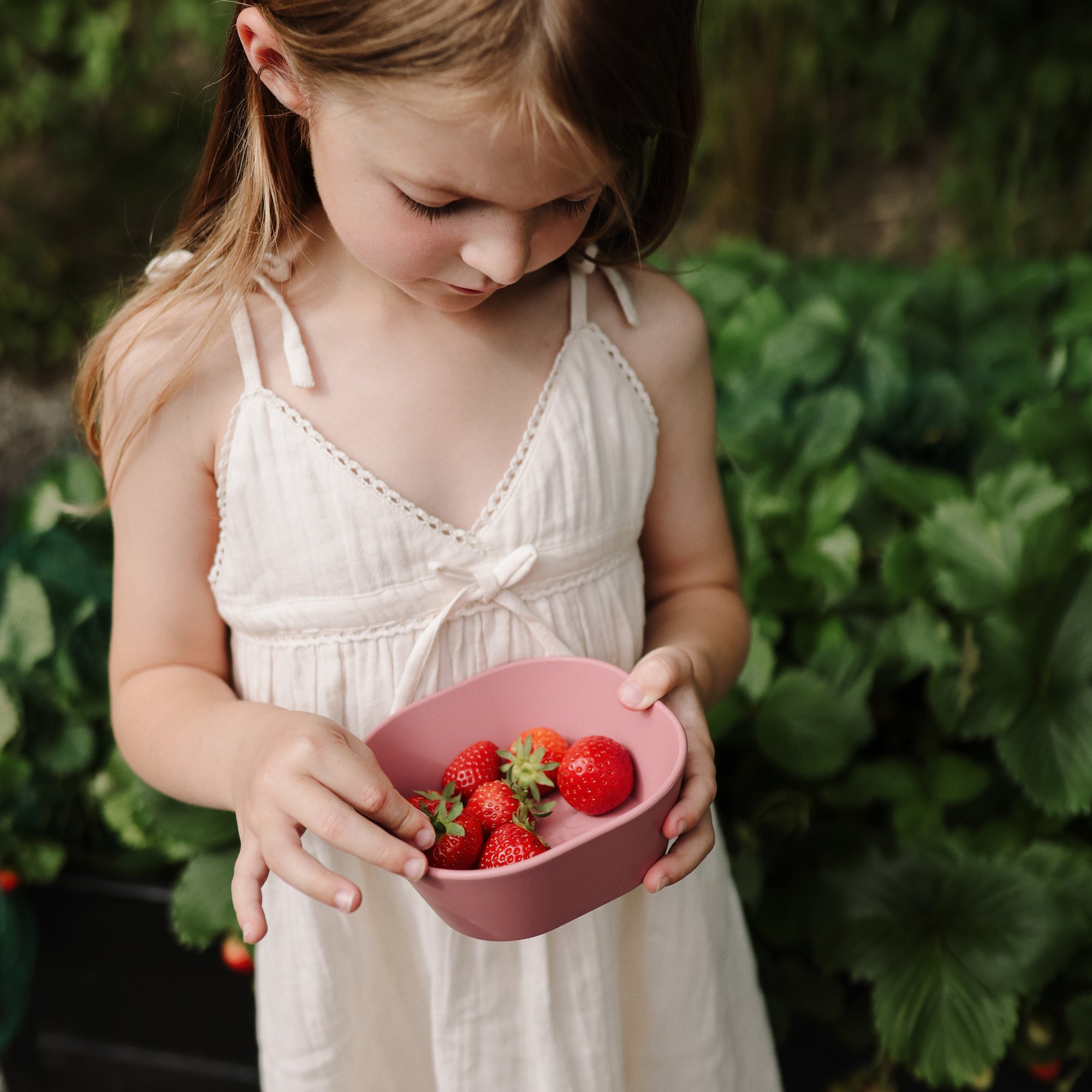 Young girl in a white dress holding mushie Square Dinnerware Bowl in a garden with strawberries.