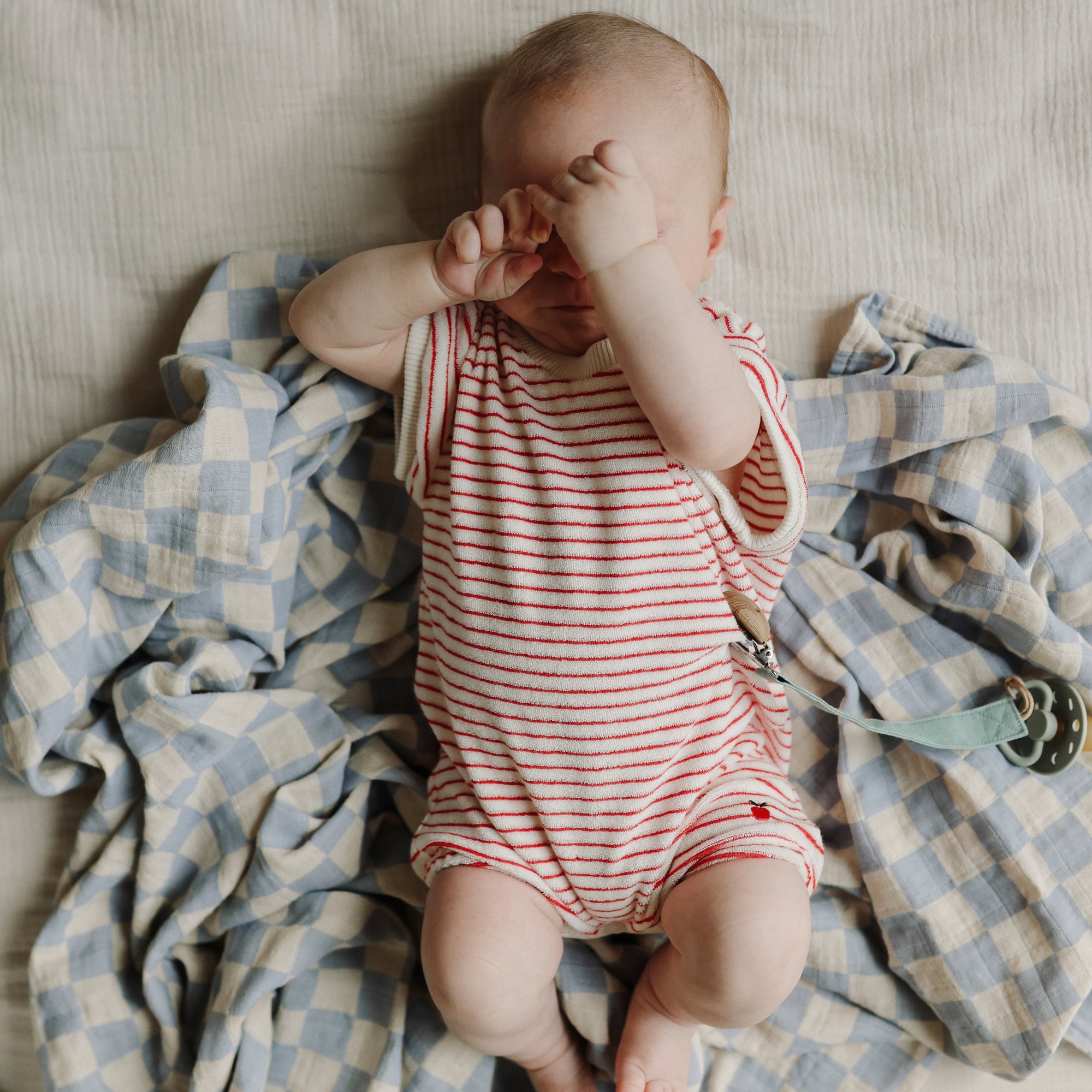 Baby lies on mushie Organic Cotton Muslin Swaddle Blanket, hands covering face.