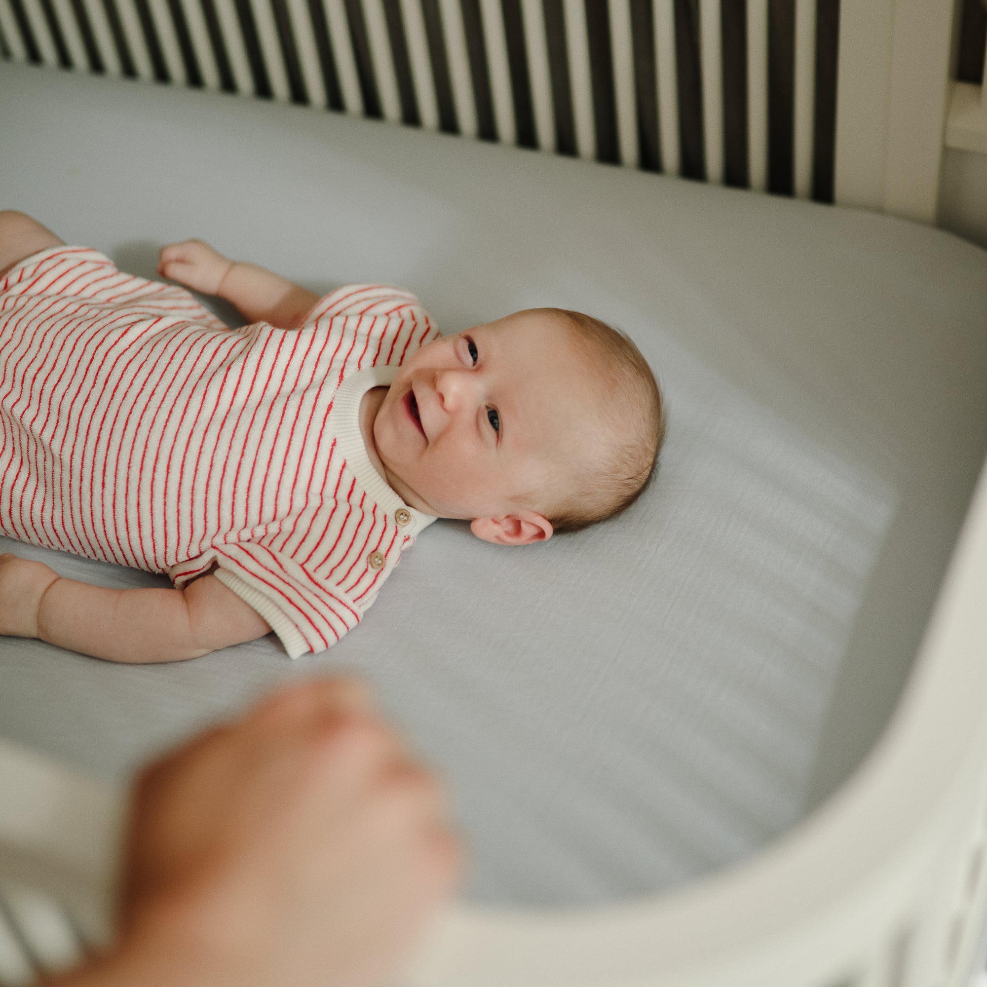 A baby in a striped onesie smiles in a crib with a mushie Extra Soft Muslin Crib Sheet.