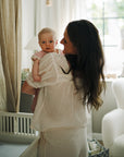 Woman holds baby in nursery with mushie Extra Soft Muslin Crib Sheet and breathable bedding.