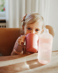 Young child uses Mushie Trainer Sippy Cup with easy-grip handles at the table.