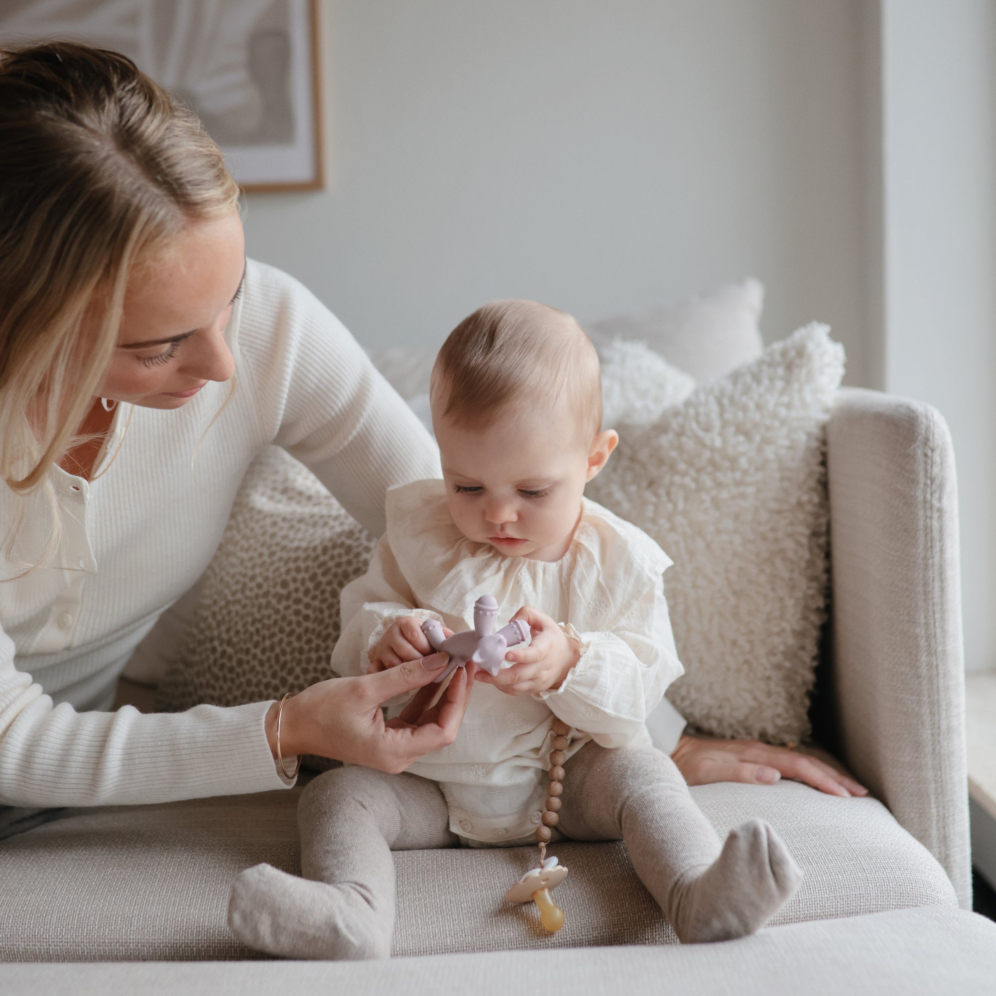 A woman and baby examine a mushie Figurine Teether together while sitting on a sofa.