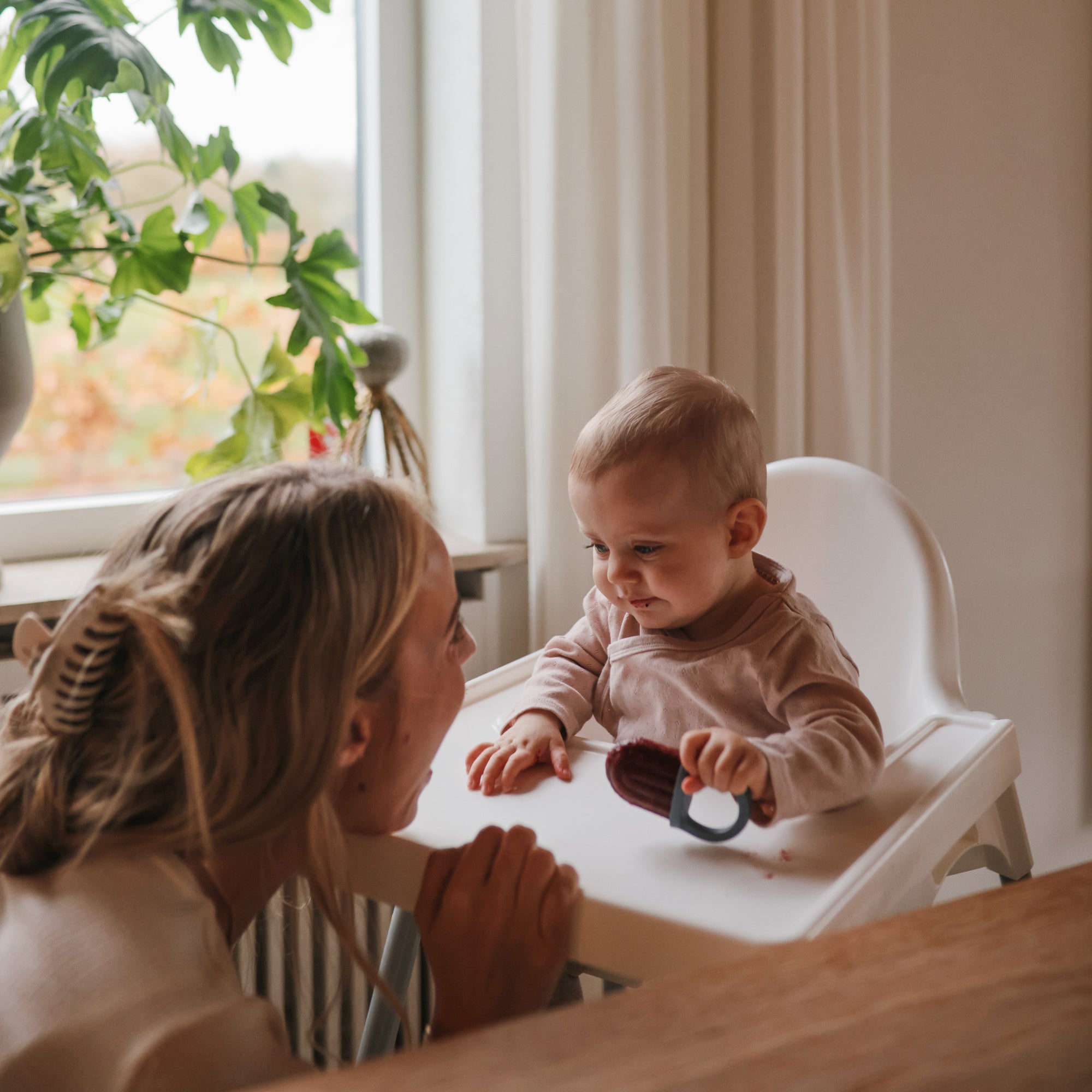 A woman smiles at a baby in a high chair holding mushie Toddler Ice Pop Tray popsicles.