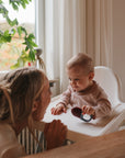 A woman smiles at a baby in a high chair holding mushie Toddler Ice Pop Tray popsicles.