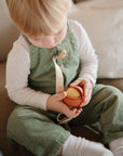 Toddler in green overalls holds mushie Nesting Spheres Sensory Toy while sitting on the couch.