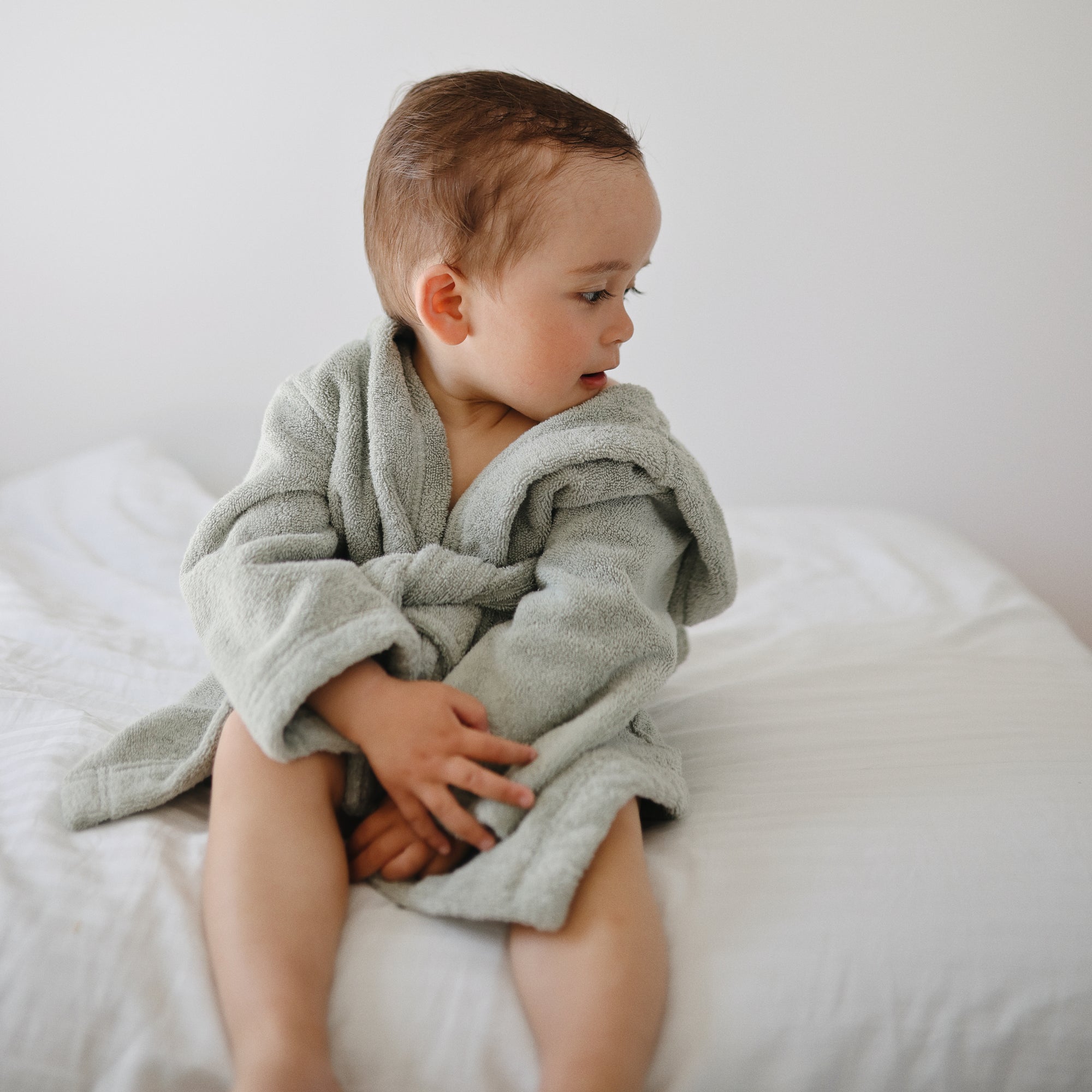 A toddler in a mushie Bear Robe sits on a bed, looking to the side against a white background.