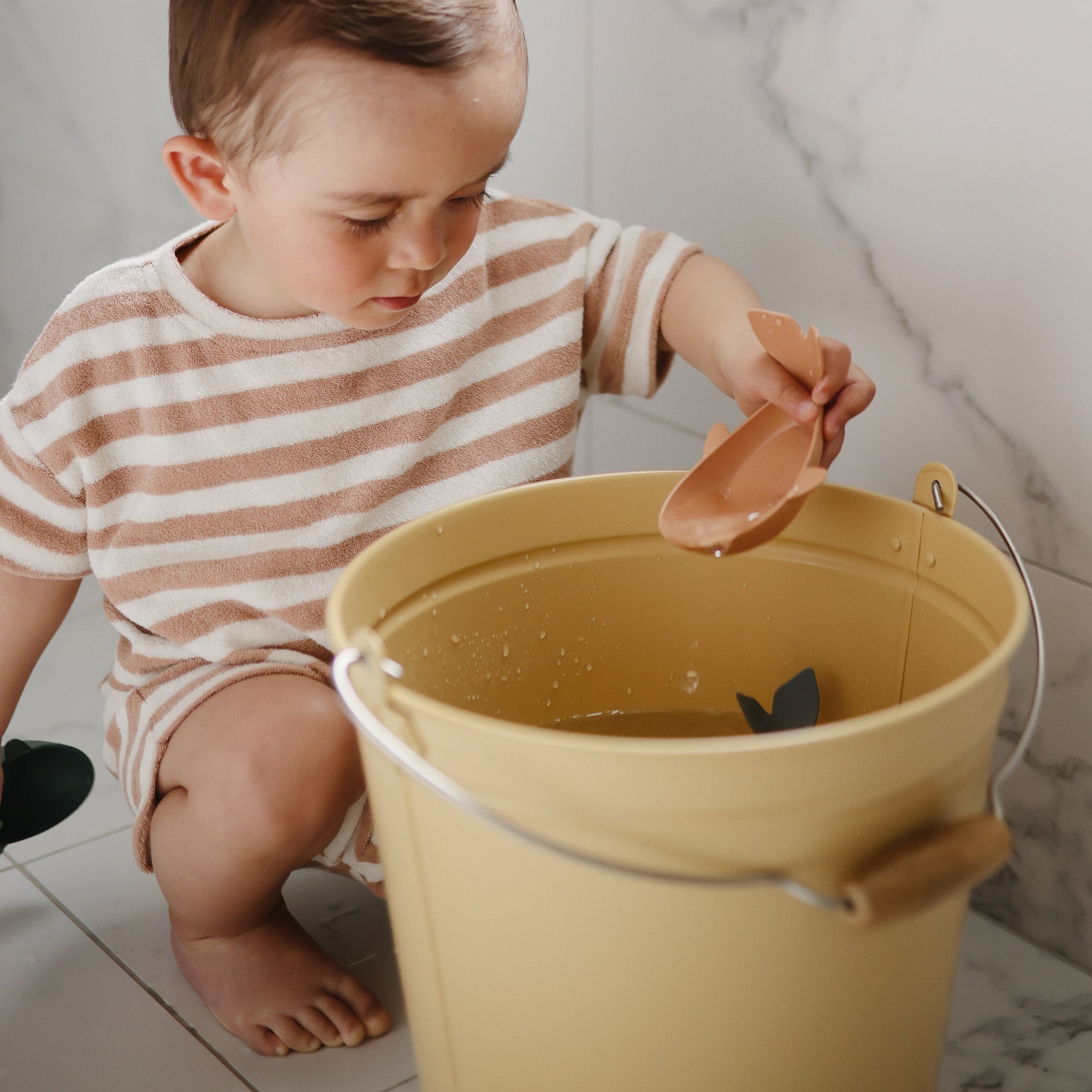 A child plays with Mushie Whale Bath Stackers, scooping water from a yellow bucket.
