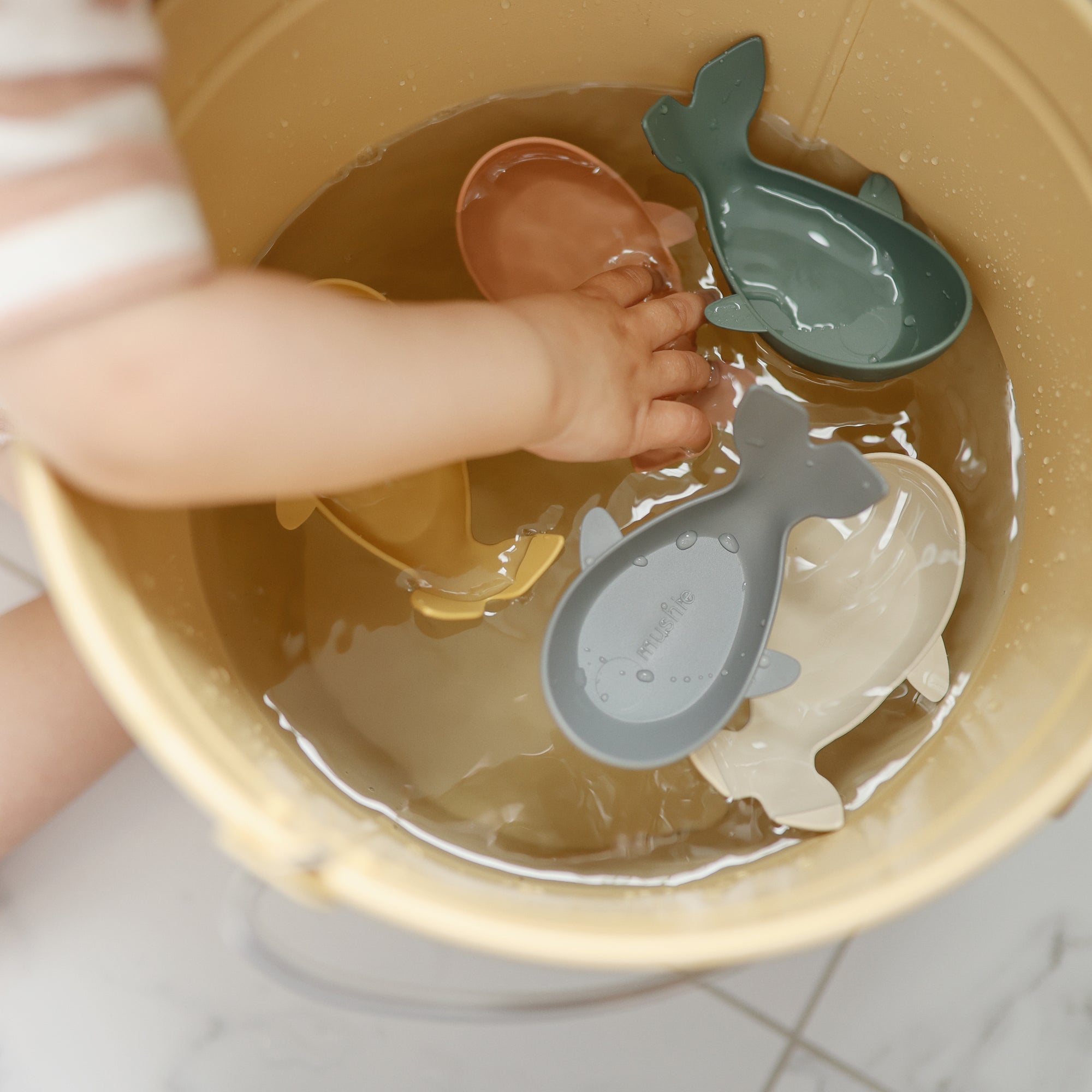Child’s hand reaching for Mushie Whale Bath Stackers and colorful bath toys in a bucket.