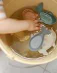 Child’s hand reaching for Mushie Whale Bath Stackers and colorful bath toys in a bucket.