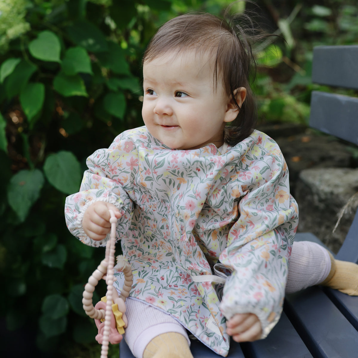 Happy baby outside in a floral mushie Long Sleeve Bib, holding a beaded toy.