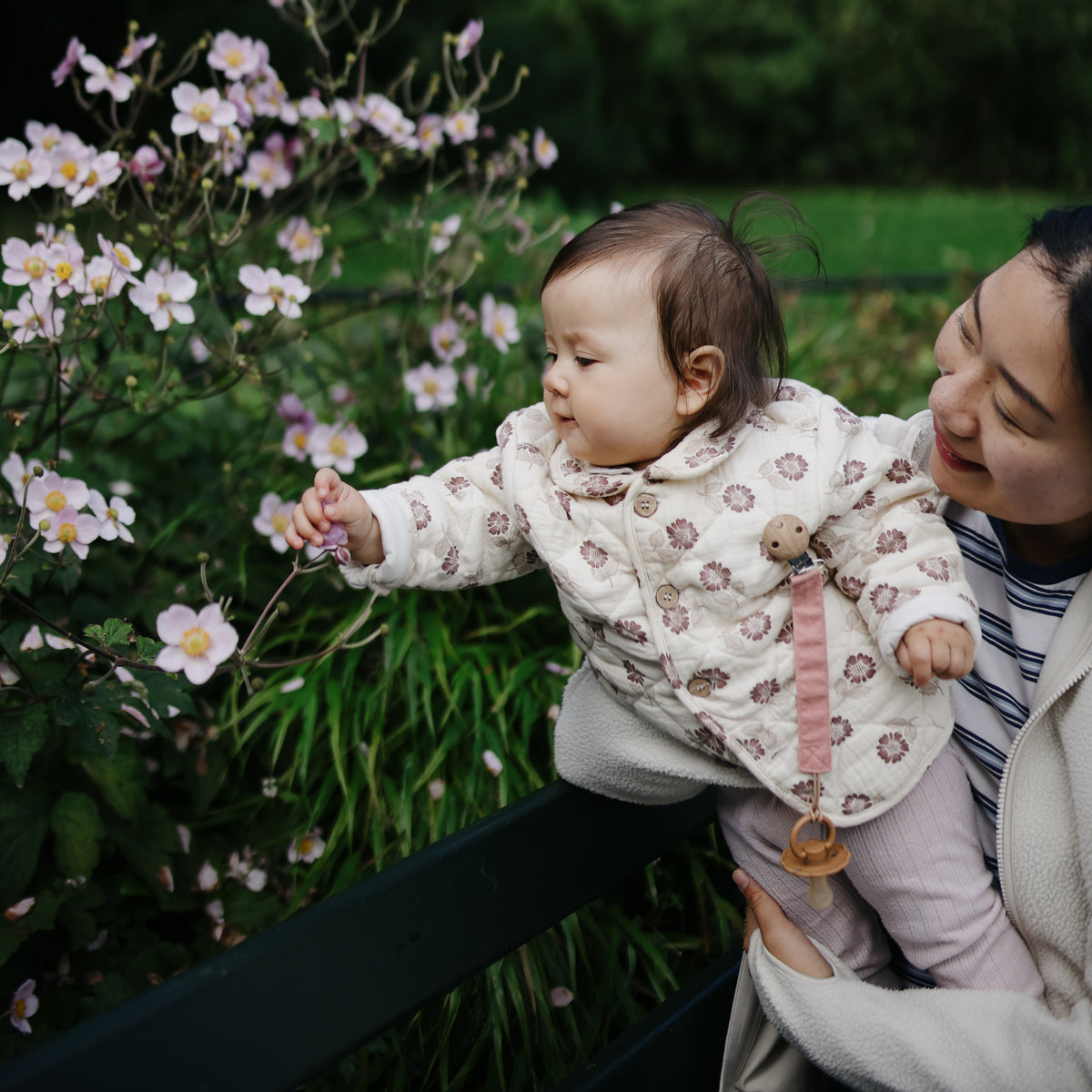 A woman holds a baby reaching for flowers, wearing the mushie Linen Pacifier Clip 2-Pack.