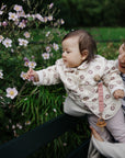 A woman holds a baby reaching for flowers, wearing the mushie Linen Pacifier Clip 2-Pack.
