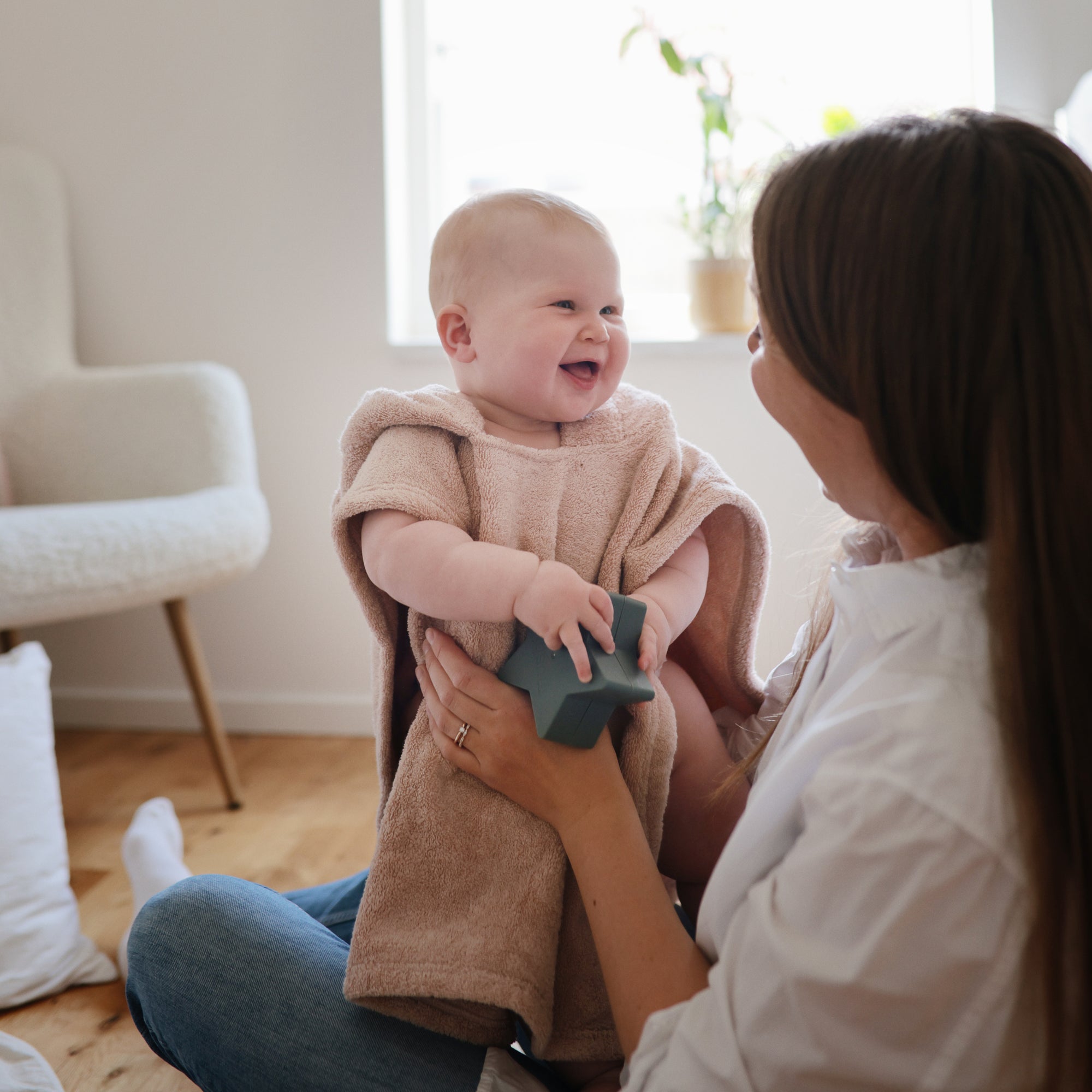 Smiling baby in mushie Bear Poncho Towel, holding a toy on woman’s lap indoors.