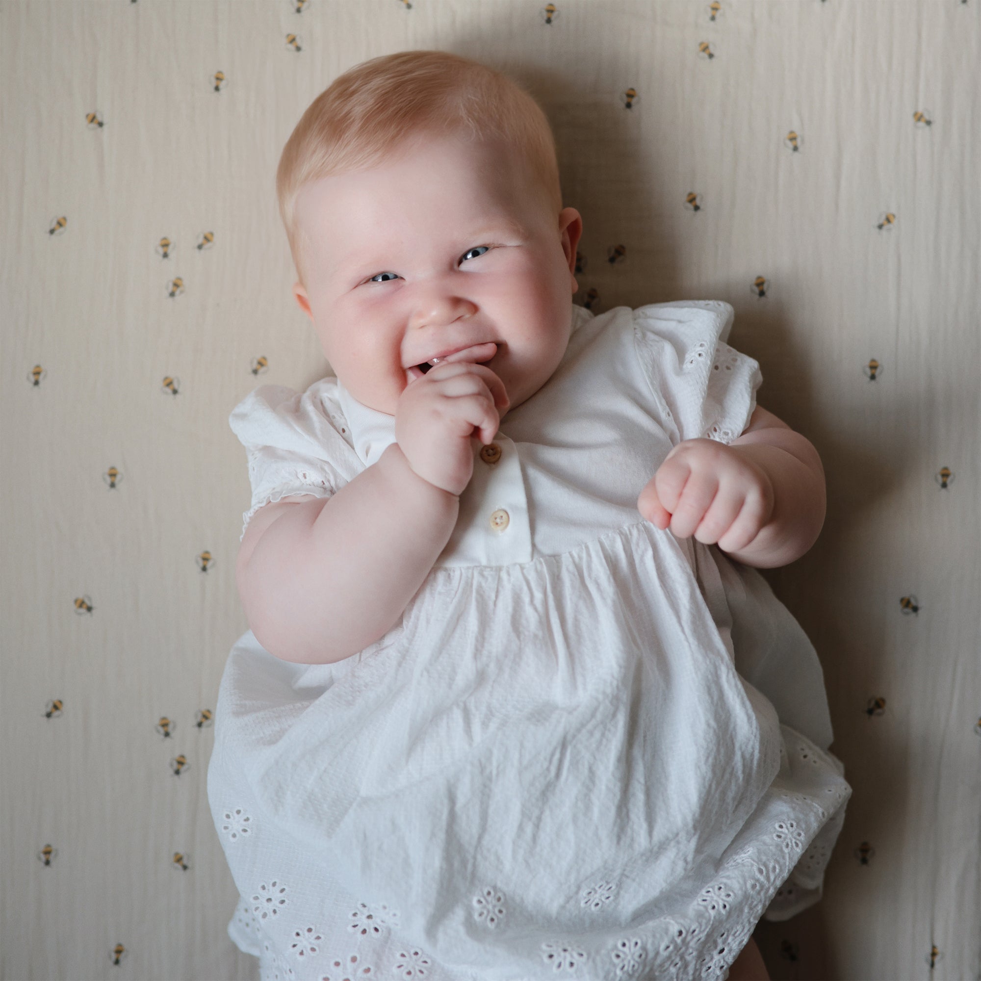 Smiling baby on mushie Extra Soft Muslin Crib Sheet, hand near mouth, wearing white dress.