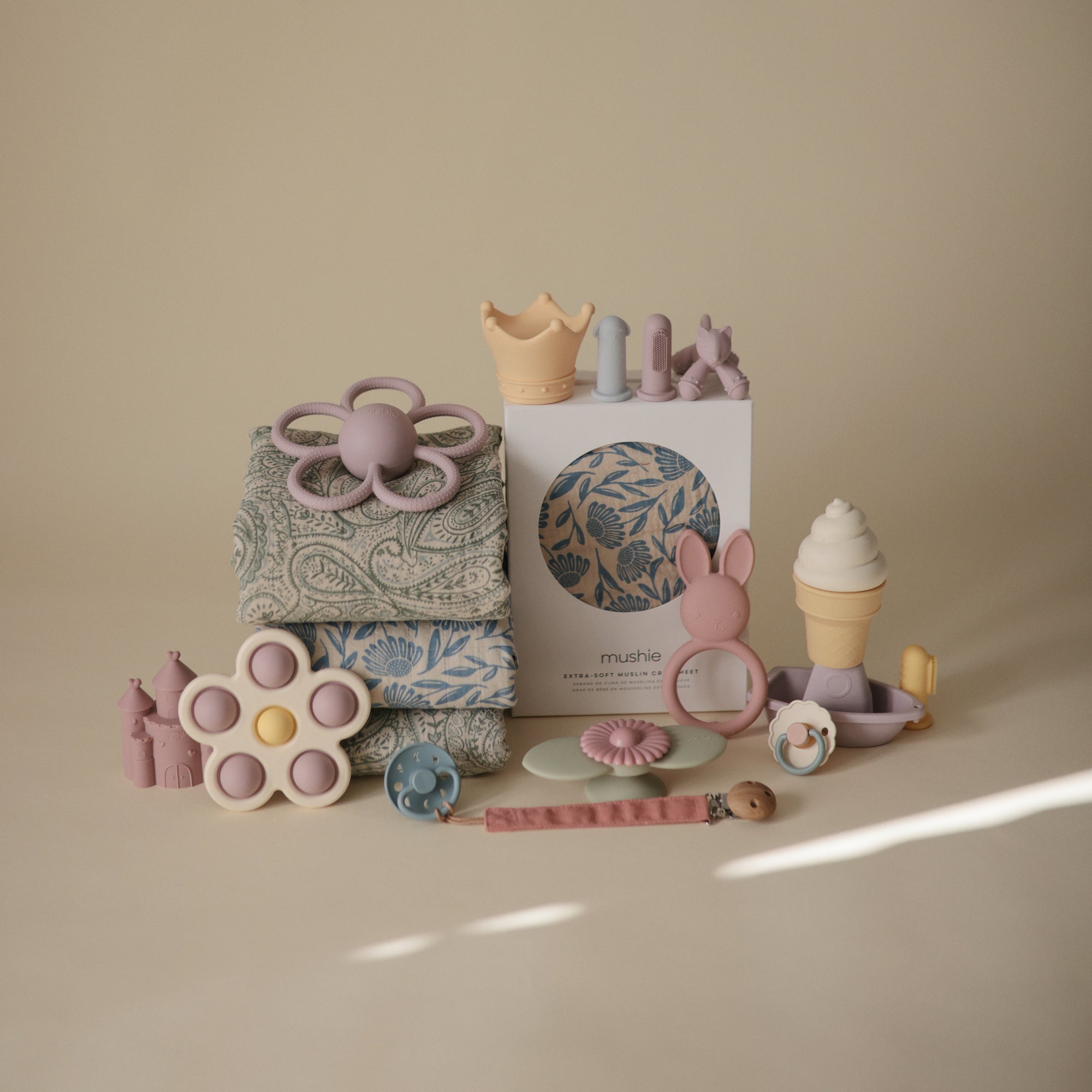 Mushie Extra Soft Muslin Crib Sheet displayed with assorted pastel baby toys on a light background.